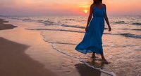 Woman in flowing blue dress walking on beach at sunset with golden sky and ocean waves creating romantic peaceful atmosphere for vacation travel concepts.