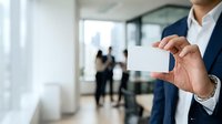 Businessman in formal navy suit and white shirt holding blank business card in modern office environment with colleagues in background and glass partitions.
