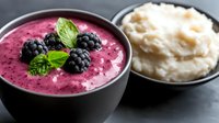 Fresh blackberry smoothie bowl topped with whole berries and mint leaves alongside creamy mashed potatoes in dark ceramic bowls on black background.