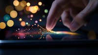 Magical glowing particles scatter from finger touch on digital screen with colorful bokeh lights in dark background, creating ethereal interactive technology effect.