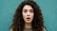 Surprised young woman with curly hair showing shocked facial expression against turquoise background for emotional reactions and lifestyle concepts.