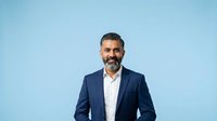Middle-aged man with dark hair and beard, appearing to be of Mediterranean or Middle Eastern ethnicity, wearing navy blazer and white shirt against blue background for corporate portraits.