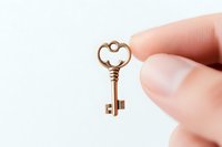 Vintage brass key with heart-shaped bow held between fingers against white background, isolated decorative element for design projects and romance concepts.