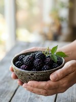 Hands holding rustic bowl filled with fresh ripe blackberries and green leaves on wooden surface, perfect for food, agriculture, harvest, and healthy eating content.