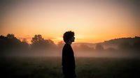Young African American man silhouette standing in misty field during golden hour sunrise with warm orange sky and peaceful morning atmosphere.