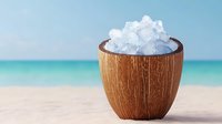 Coconut shell bowl filled with ice cubes on tropical beach with turquoise ocean and white sand background for summer refreshment concepts.