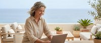 Middle-aged woman with blonde hair working on laptop at coastal terrace with ocean view, potted plants, and comfortable seating for remote work or business concepts.
