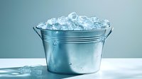 Metal bucket filled with crystal clear ice cubes on white surface against blue background, cold refreshing drops of condensation on shiny steel surface.