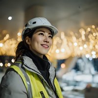 Asian female construction worker in hard hat and safety vest smiling at industrial site with warm bokeh lights creating inspiring workplace atmosphere.