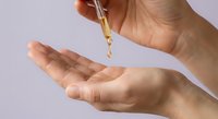 Woman applying golden serum oil drops from pipette dropper onto palm for skincare routine and beauty treatment on light background.