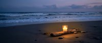 Glowing candle on driftwood creates warm ambiance on sandy beach at twilight with ocean waves and purple sky for romantic evening atmosphere.