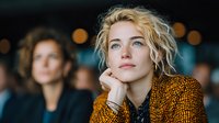Young blonde businesswoman in golden sequined blazer contemplating future opportunities during corporate evening event with colleagues in background.