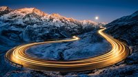 Winding mountain road with golden light trails from vehicle headlights at dusk, snow-covered alpine peaks under moonlit sky, long exposure night photography.