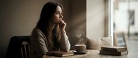 Thoughtful young woman with long dark hair sitting at wooden table with coffee cup and books, contemplating by window in warm natural light.