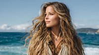 Young woman with long wavy blonde hair gazing at ocean horizon, wearing bohemian style top against turquoise sea background. Perfect for travel and lifestyle content.