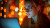 Young Caucasian woman in glasses illuminated by warm orange bokeh lights works on laptop in evening. Side profile portrait shows contemplative expression and cozy sweater.