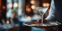 Elegant martini cocktail with sugar rim on silver tray served by waiter in upscale restaurant with atmospheric bokeh lighting.