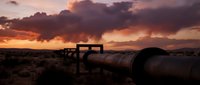 Industrial pipeline infrastructure stretching across desert landscape during dramatic sunset with stormy clouds and orange sky creating moody atmosphere.