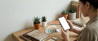 Female graphic designer working at wooden desk with smartphone, color palette samples, and small potted plants in modern home office workspace.
