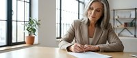 Professional businesswoman in beige suit writing at modern office desk with natural lighting from large windows and minimalist workspace design.