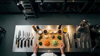 Professional chef preparing colorful vegetable ingredients on cutting board in commercial kitchen with knives and cooking utensils overhead view.
