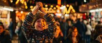 Young Asian boy making heart shape with hands at festive night market with warm bokeh lights creating joyful holiday atmosphere for family celebrations.