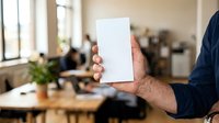 Man holding blank white card in modern office workspace with colleagues working at desk in background.