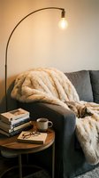 Cozy reading nook with gray upholstered armchair, cream knit throw blanket, wooden side table with stacked books, eyeglasses, coffee mug, and arched floor lamp with warm lighting for relaxation and home decor.