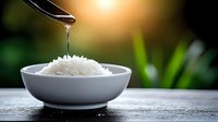 Coconut oil dripping from spoon onto white rice in ceramic bowl on wooden table with tropical plants background and warm sunlight.