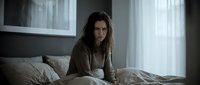 Young woman with long dark hair and olive complexion sitting on bed in modern bedroom with natural light from windows, expressing concern or sadness, suitable for mental health, wellness, lifestyle and emotional content.