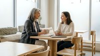 Two professional women having business meeting at wooden table in modern office with natural light discussing strategy and collaboration.