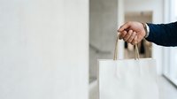 Close-up of hand holding white shopping bag with handles in modern interior space, suitable for retail, commerce, and lifestyle marketing campaigns.