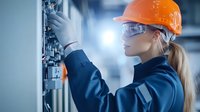 Female industrial electrician in protective gear working on electrical cabinet, wearing orange hard hat and safety glasses, adjusting control panel in modern facility.