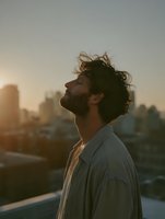 Middle Eastern man with curly hair and beard in casual white shirt standing on rooftop at sunset, city skyline silhouette in background, atmospheric golden hour lighting.