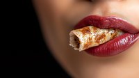 Sensual red lips holding crispy rolled wafer dessert against dark background, close up macro shot of mouth with burgundy lipstick and sweet treat.