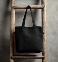 Black canvas tote bag hanging on rustic wooden ladder against concrete wall. Minimalist eco-friendly shopping accessory for everyday use.