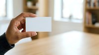 Close-up of hand holding blank white business card in modern office interior with wooden desk and blurred background for branding mockup.
