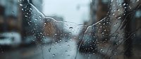 Raindrops on car windshield with wipers during storm creating abstract water patterns and blurred urban background for weather concepts.