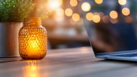 Glowing mason jar candle holder on wooden desk with laptop and plant, warm bokeh lights creating cozy atmosphere for evening work or relaxation.