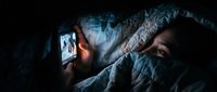 Young woman lying in bed at night using smartphone under blanket in dark bedroom creating cozy atmosphere for late evening social media browsing.