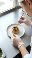 Chef in white gloves plating gourmet appetizer with tweezers on white ceramic dish near window. Fine dining food preparation and culinary artistry.