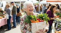 Happy senior Caucasian woman holding fresh vegetables at farmers market. Elderly lady with white hair smiling while shopping for organic produce outdoors.