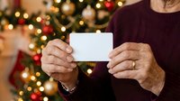 Senior man holding blank white card with festive holiday decorations and twinkling lights in background, suitable for greeting cards, invitations, promotions, and seasonal marketing campaigns.