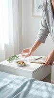 Woman in linen shirt reaching for glass of water on white bedside table with notebook and healthy snacks in bright minimalist bedroom interior.