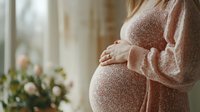 Young pregnant woman in cozy knit sweater gently holding baby bump near window with soft natural light and pink roses, capturing tender maternal moment.
