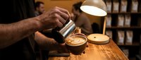 Skilled barista pouring steamed milk into a latte art cup at a cozy coffee shop counter with warm lamp light in the background.