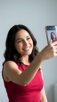 Happy young Hispanic woman taking selfie with smartphone wearing red top smiling at camera for social media content creation and digital communication.