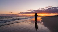 Silhouetted runner jogging on wet beach sand during golden sunset with ocean waves and dramatic sky, suitable for fitness and wellness marketing.