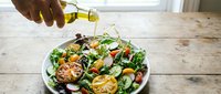 Drizzling olive oil over fresh salad with grilled vegetables, lemon slices, and greens on wooden table for healthy eating and cooking.