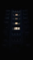 Illuminated windows in dark apartment building create mysterious atmosphere. Glowing lights reveal vertical pattern of residential structure at night.